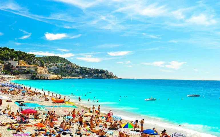 A popular Mediterranean beach filled with people and brightly coloured beach umbrellas with a clear blue sea and clear blue sky.